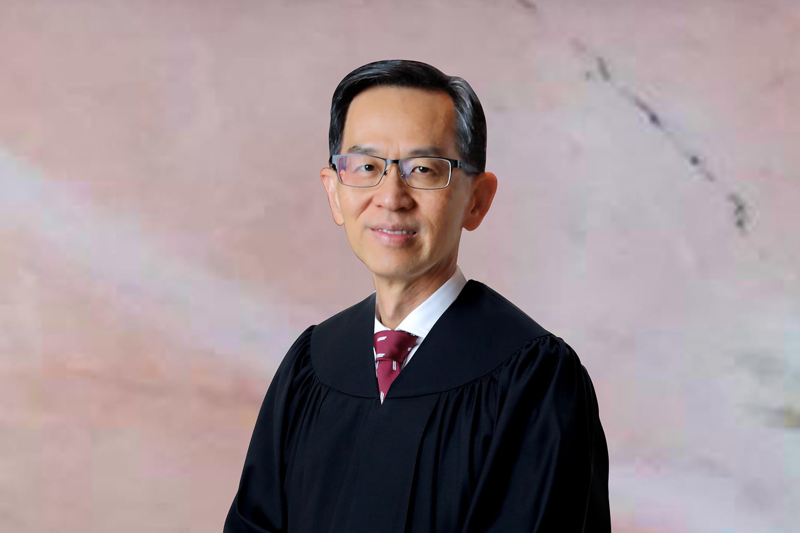 Head and shoulders of man in black judge's robe, glasses, maroon tie against mottled background.